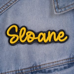 May include: A yellow embroidered patch with the name "Sloane" in a cursive font, outlined in black. The patch is set against a light blue denim background.