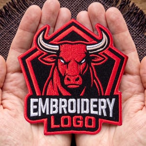May include: A pentagonal embroidered patch featuring a red bull head with white horns and eyes set against a black background. The patch is outlined in red and includes the white text EMBROIDERY and the red text LOGO at the bottom section.