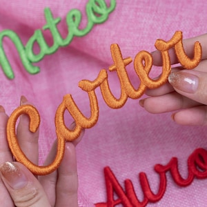 May include: Embroidered name patches in orange, green, and red script lettering. The orange "Carter" patch is held by a person. The green "Nate" and red "Auro" patches are also visible. The patches are on a pink background.