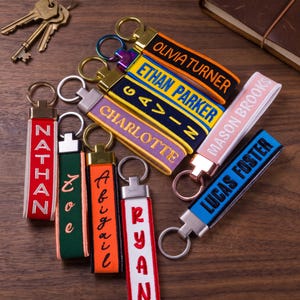 May include: A collection of personalized fabric keychains featuring various colors including red, green, orange, white, blue, and purple. Each keychain has a metal ring and is embroidered with names such as Olivia Turner, Ethan Parker, Gavin, Charlotte, Mason Brooks, Lucas Foster, Nathan, Zoe, Abigail, and Ryan.