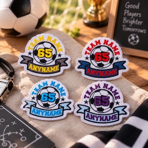 May include: Four embroidered soccer patches featuring a central soccer ball graphic with the number 65 in the center. Each patch includes the text TEAM NAME arched above and ANYNAME on a banner below, available in yellow, red, blue, and purple.