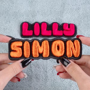 May include: Two embroidered name patches. One patch reads "LILLY" in bright pink thread on a black background. The other patch reads "SIMON" in orange thread on a black background. Both patches have a wavy black border.