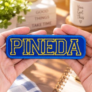 May include: A rectangular embroidered patch with a royal blue background and a thick royal blue border. The word PINEDA is stitched in the center in a bold, yellow, collegiate-style font with a thin yellow outline around each letter.