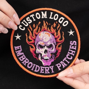 May include: Embroidered patch with a skull design. The skull is purple and white, surrounded by orange and red flames. The patch has the text "CUSTOM LOGO" and "EMBROIDERY PATCHES" in a circular design, with two white stars. The patch has a black background and an orange border.