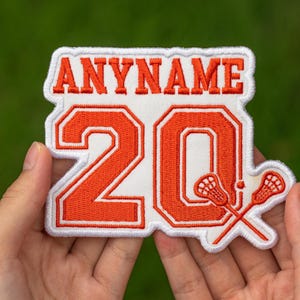 May include: An orange and white embroidered patch with the text "ANYNAME" above the number "20". The patch features a white border and crossed lacrosse sticks. Ideal for sports apparel or accessories.