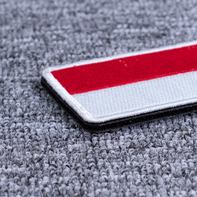 Pode incluir: Remendo retangular com um design listrado vermelho e branco, contornado por uma borda preta. O remendo &eacute; exibido em um fundo cinza texturizado, destacando as cores vibrantes e as costuras detalhadas.