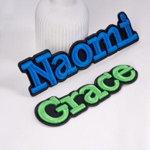 May include: Two embroidered name patches. One patch reads "Naomi" in blue letters with a black border. The other patch reads "Grace" in green letters with a black border. Both patches are on a white surface.