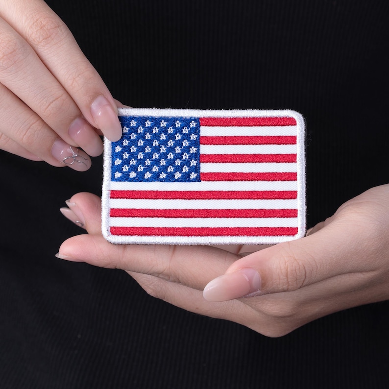 Pode incluir: Um patch retangular com o design da bandeira americana. O patch tem uma borda branca, um campo azul com estrelas brancas e listras vermelhas e brancas. O patch &eacute; segurado nas m&atilde;os de uma pessoa contra um fundo preto.