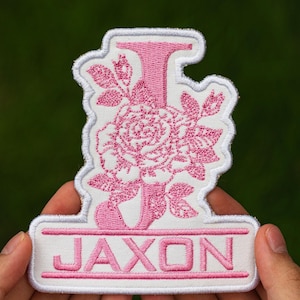 May include: Embroidered patch featuring the name "JAXON" in pink, with a pink floral design and the letter "J". The patch has a white background and a white border. The patch is held in a person's hands.