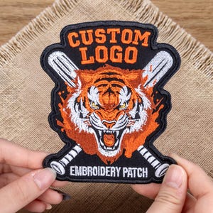 May include: A black embroidered patch featuring an orange tiger head in the center, flanked by two crossed white baseball bats. The text CUSTOM LOGO is stitched in orange at the top, and EMBROIDERY PATCH is stitched in white at the bottom.