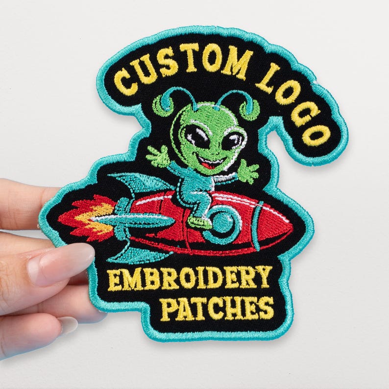 May include: Embroidered patch featuring a green alien riding a red rocket. The patch has a black background with the words "CUSTOM LOGO" and "EMBROIDERY PATCHES" in yellow. The patch is outlined in teal.