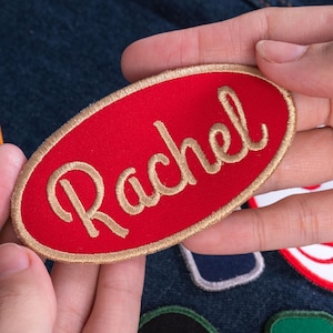 May include: An oval-shaped embroidered patch with a vibrant red background and a gold-colored border. The name Rachel is stitched in the center in a gold-colored cursive script font. The patch is designed for attachment to fabric items.