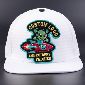 May include: White baseball cap with a flat brim, featuring a colorful embroidered patch. The patch depicts a green alien riding a red rocket, with the text "CUSTOM LOGO EMBROIDERY PATCHES" in a curved design.