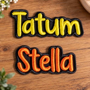 May include: Two embroidered name patches with black fabric backgrounds. The top patch features the name Tatum in bright yellow thread, while the bottom patch displays the name Stella in vibrant orange thread. Both patches have a scalloped edge.