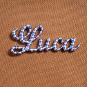 May include: Embroidered name "Luca" in a cursive script, featuring blue and purple stripes on a white background. The stitching is on a textured, brown fabric.