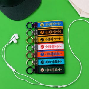 May include: A collection of eight fabric keychains featuring embroidered sound wave patterns and musical note icons in various colors including blue, black, yellow, white, orange, light blue, and dark green, each attached to a metal ring.