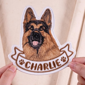 May include: An embroidered patch featuring a German Shepherd dog head with brown and black fur, a black nose, and a pink tongue. Below the dog is a white banner with brown borders containing the name CHARLIE and two brown paw prints.