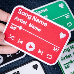 May include: A rectangular red embroidered patch featuring a white music player interface design. The text reads Song Name and Artist Name, with a progress bar, play button, skip icons, a heart symbol, and time stamps 0:37 and 3:17 in white thread.