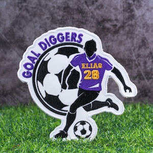 May include: A soccer-themed patch featuring a player kicking a soccer ball. The patch has the words "GOAL DIGGERS" in purple and white. The player is wearing a purple jersey with the name "ELIAS" and the number "28" in gold.