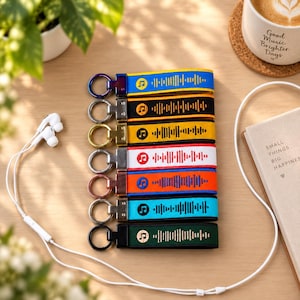 May include: A set of seven fabric keychains arranged vertically, each featuring a unique color, a metal ring, and a printed sound wave graphic with a musical note icon. The colors include blue, black, yellow, white, orange, light blue, and dark green.