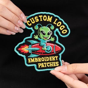 May include: Embroidered patch featuring a green alien riding a red rocket. The patch has a teal border and includes the text "CUSTOM LOGO" and "EMBROIDERY PATCHES" in yellow. The alien is smiling and wearing a teal outfit.