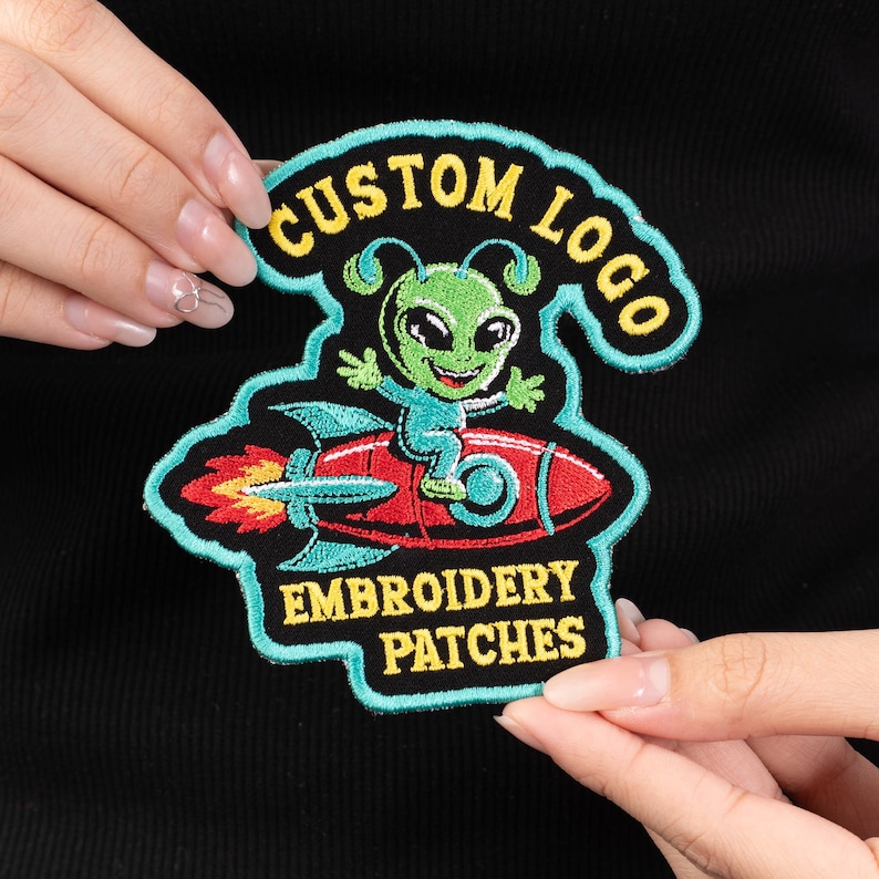 May include: Embroidered patch featuring a green alien riding a red rocket. The patch has a teal border and includes the text "CUSTOM LOGO" and "EMBROIDERY PATCHES" in yellow. The alien is smiling and wearing a teal outfit.