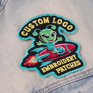 May include: Embroidered patch featuring a green alien riding a red rocket. The patch has a black background with the words "CUSTOM LOGO" and "EMBROIDERY PATCHES" in yellow. The patch is outlined in teal.