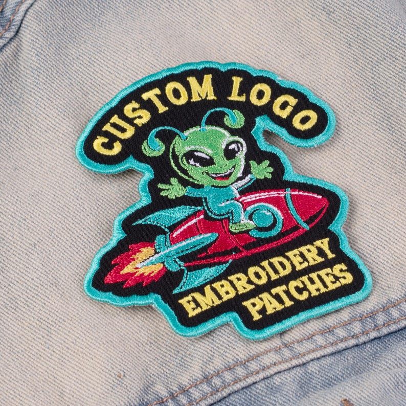May include: Embroidered patch featuring a green alien riding a red rocket. The patch has a black background with the words "CUSTOM LOGO" and "EMBROIDERY PATCHES" in yellow. The patch is outlined in teal.