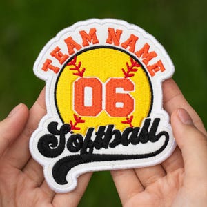 May include: Softball team patch with a yellow softball design, featuring the number 06 in orange. The patch has the words "TEAM NAME" arched across the top in orange and "Softball" in black script at the bottom. The patch is white with orange and black accents.