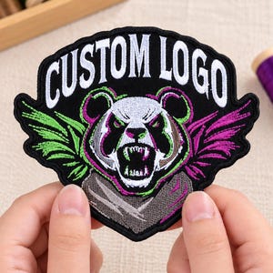 May include: A black embroidered patch featuring a snarling panda head with green and purple accents, wings on either side, and the text CUSTOM LOGO in white capital letters arched across the top of the shield-shaped design.