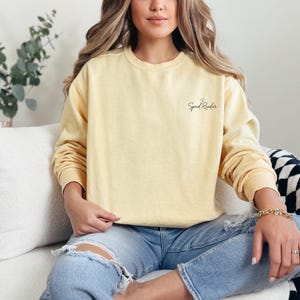 Può includere: Una felpa giallo pallido a maniche lunghe. Il testo "Speed Reader" è scritto in una sceneggiatura delicata sulla parte superiore del petto. La felpa è abbinata a jeans blu.