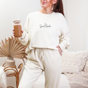 Può includere: Completo di felpa e pantaloni della tuta color crema. La felpa presenta la scritta "Speed Reader" con un piccolo cuore sopra. Un bicchiere trasparente con una bevanda marrone e una cannuccia è tenuto in mano.