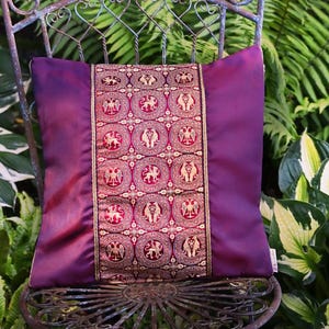 Könnte beinhalten: Ein dekoratives lila Kissen mit einem gold-rot gemusterten Mittelteil, das auf einem Vintage-Metallstuhl liegt. Das Design des Kissens zeigt sich wiederholende kreisförmige Motive mit Tierfiguren. Der Stuhl steht vor einem Hintergrund aus üppigem grünem Laub.