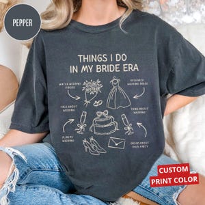 Puede incluir: Camiseta gris oscuro con el texto "THINGS I DO IN MY BRIDE ERA" e ilustraciones relacionadas con bodas. Incluye opción de color de impresión personalizado.