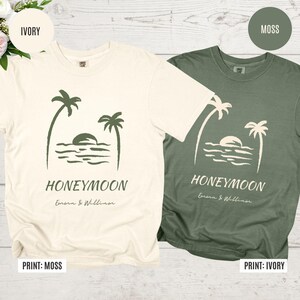 May include: Two t-shirts, one ivory and one moss green, featuring a palm tree and sunset design. The word "HONEYMOON" is printed below the graphic, along with the names "Emma & William".