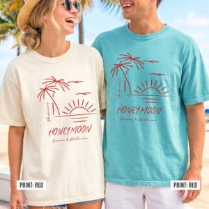 May include: Two t-shirts, one cream and one teal, featuring a red graphic of palm trees, a sunset, and the word "Honeymoon." The text "Hawaii" and names are also included. The shirts are casual and suitable for a vacation theme.
