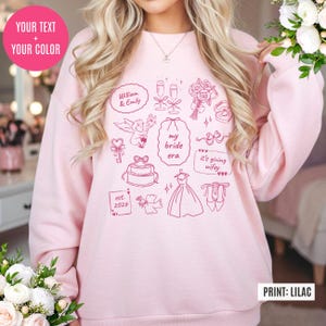 May include: A light pink sweatshirt with a variety of pink illustrations related to weddings, including champagne glasses, a cake, and a wedding dress. Text includes "William & Emily", "my bride era", and "est. 2026". The print color is lilac.