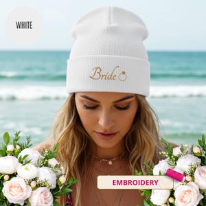 May include: White beanie with the word "Bride" embroidered in gold script, along with a ring design. The hat is ribbed and has a folded cuff. The word "WHITE" is in a white circle in the upper left corner. The word "EMBROIDERY" is in a pink rectangle.