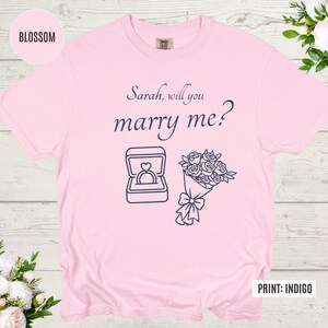 May include: A light pink t-shirt with the text "Sarah, will you marry me?" in navy blue script. Below the text are navy blue line drawings of a ring in a box and a bouquet of flowers. The shirt is a Comfort Colors brand.