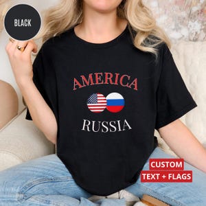 May include: Black t-shirt featuring the words "AMERICA" and "RUSSIA" in red and white. The design incorporates the American and Russian flags. The shirt also has the text "CUSTOM TEXT + FLAGS".