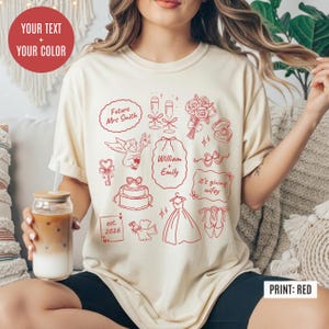 May include: Cream-colored t-shirt with red wedding-themed graphics, including text that says "Future Mrs Smith", "William + Emily", and "it's giving wifey". The shirt also features a wedding cake, dress, and champagne glasses.