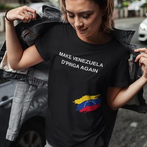 May include: Black t-shirt with the text "MAKE VENEZUELA D'PINGA AGAIN" in white. The shirt features a Venezuelan flag design in yellow, blue, and red. The shirt is worn by a person in a denim jacket and shorts.