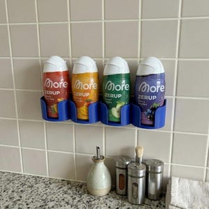 Op de afbeelding: Vier flessen ZERUP-siroop in rood, geel, groen en paars, elk in een blauwe houder. De flessen zijn gelabeld met de smaken: Perzik & Thee, Mango Orange, Groene Appel en Bessen Medley. De flessen zijn op een betegelde muur gemonteerd.