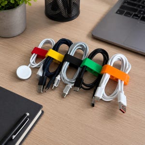 Könnte beinhalten: Fünf aufgewickelte Ladekabel, die mit bunten Silikon-Kabelbindern in Rot, Gelb, Schwarz, Grün und Orange befestigt sind. Die Kabel umfassen ein weißes magnetisches Uhrenladegerät, ein schwarzes geflochtenes Kabel, ein silbernes geflochtenes Kabel und zwei weiße Kabel.