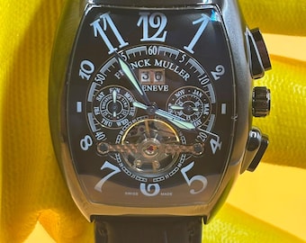 Könnte beinhalten: Eine schwarze Franck Muller Geneve Armbanduhr mit tonnenförmigem Gehäuse und schwarzem Lederarmband. Das schwarze Zifferblatt zeigt große weiße arabische Ziffern, ein Datumsfenster, mehrere Hilfszifferblätter für Tag und Monat sowie ein Tourbillon.
