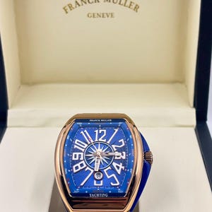 Könnte beinhalten: Eine Franck Muller Geneve Armbanduhr mit einem roségoldfarbenen Tonneau-Gehäuse und einem blauen Zifferblatt mit Kompassrosen-Design, großen weißen arabischen Ziffern und dem Schriftzug YACHTING. Die Uhr hat ein blaues Armband und liegt in einer Box.