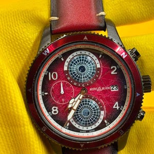 Könnte beinhalten: Eine Montblanc Armbanduhr mit tiefrotem Farbverlaufszifferblatt, zwei Hilfszifferblättern mit Globusmuster, weißen arabischen Ziffern bei 2, 4, 8 und 10, Datumsanzeige bei 3 Uhr und einer roten drehbaren Lünette mit Himmelsrichtungen am Lederarmband.