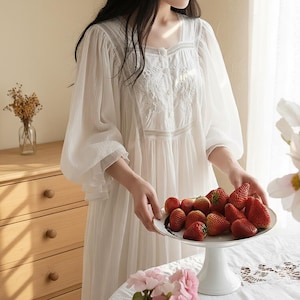 Puede incluir: Un camisón blanco de manga larga con detalles bordados alrededor del escote y el corpiño. El camisón tiene un escote cuadrado y cierre de botones. Se sostiene un plato de fresas frescas.