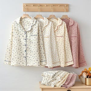 Op de afbeelding: Vier sets pyjama's in crème, geel en roze, elk met een overhemd met knopen en een bijpassende broek. De crèmekleurige set heeft een blauw bloemenpatroon, de gele heeft gouden stippen en de roze heeft een bloemenstructuur. De pyjama's hangen aan houten hangers.
