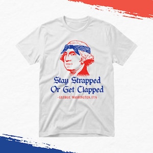 Könnte beinhalten: Ein weißes kurzärmeliges T-Shirt mit einer roten Grafik von George Washington, der ein blaues Bandana trägt. Unter der Grafik steht der Text: Stay Strapped Or Get Clapped -GEORGE WASHINGTON, 1776 in blauer und roter Schrift.